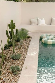 a small pool surrounded by rocks and plants with pillows on the edge, in front of a white wall