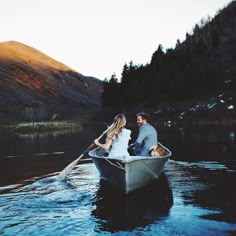 a man and woman in a row boat on the water with mountains in the background