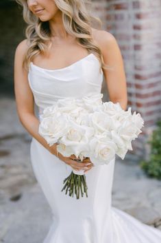 a woman in a wedding dress holding a bouquet of white roses and looking at the camera