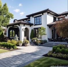 a large white house with black trim and lots of plants in the front yard, on a sunny day