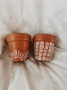 two clay pots with designs on them sitting on a white bed sheet in front of a pillow