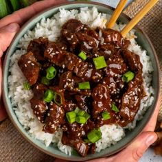 two hands holding a bowl filled with rice and beef on top of green onions, as well as chopsticks