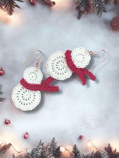 two crocheted snowman earrings with red ribbon on them and pine trees in the background