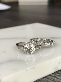 two engagement rings sitting on top of a white marble slab, with one diamond in the middle