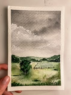 a hand holding up a piece of watercolor paper with a painting of a field in the background