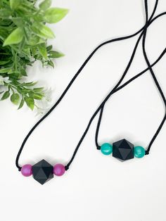 two necklaces with black and pink beads are sitting on a white surface next to a plant