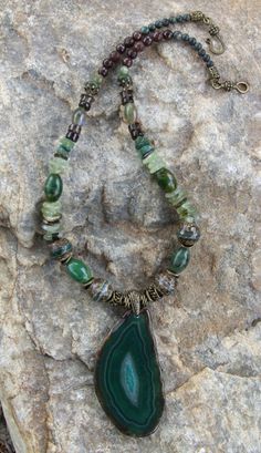 This attractive necklace is composed of many types of stones and beads:  there are 4 large tibetan line agate beads; 4 large green oval jasper gemstone beads; prehnite large chip/disc gemstone beads; fancy jasper beads; serpentine jasper beads; brown jade beads; Smokey Quartz beads; along with bronze pewter spacer beads and a hook claps, all of which are lead and nickel free. The pendant is 3 inches long and 1 1/2 inches wide, with crystals in the middle, framed by dark and light swirls of green and a few brown swirl lines.  The necklace is 20 1/4 inches long. Luxury Agate Beaded Necklace With Natural Stones, Luxury Long Agate Necklace, Luxury Green Agate Beaded Necklaces, Agate Necklace Green, Luxury Beaded Agate Necklaces, Bohemian Beaded Necklaces With Malachite Stones, Bohemian Beaded Necklace With Malachite Stones, Bohemian Beaded Malachite Necklace With Natural Stones, Healing Natural Jade Gemstones