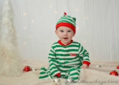 a baby wearing a green and white striped outfit sitting in front of a christmas tree