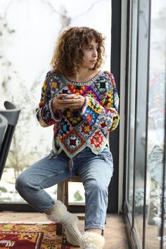 a woman sitting on a chair looking at her cell phone while wearing a multicolored crocheted sweater