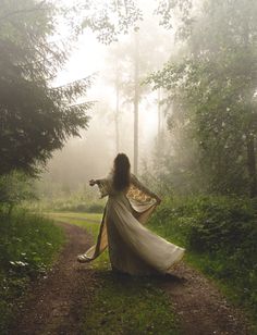 a woman in a white dress is walking through the woods on a foggy day