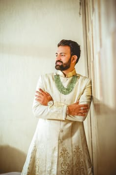 a man with a beard wearing a white suit and green necklace standing in front of a wall