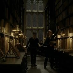 two people walking through a library with bookshelves and lamps on either side of them