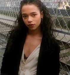 a woman with long hair standing in front of a chain link fence and looking at the camera