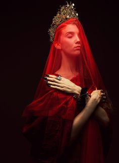 a woman wearing a red veil and ring with her hands on her chest, in front of a black background