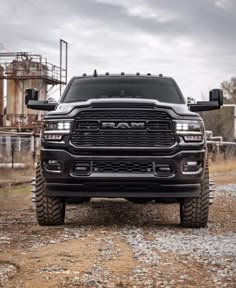 the front end of a black truck parked on top of a dirt road next to a factory