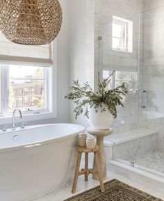 a white bath tub sitting under a window next to a wooden stool and plant in a vase