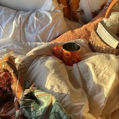 an open book is laying on the bed with blankets and pillows, next to a cup