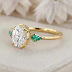 a close up of a diamond and emerald ring on a white surface with flowers in the background