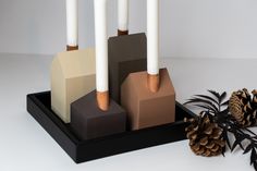 three candles are placed in different shapes and sizes on a black tray next to pine cones