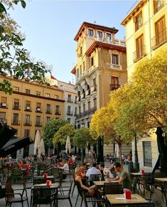 people are sitting at tables in an outdoor cafe area with tall buildings and trees on either side