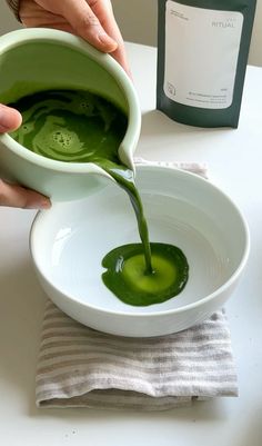 a person pouring green liquid into a white bowl on top of a table next to a bottle