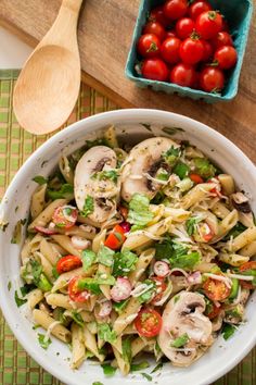 a white bowl filled with pasta salad next to a wooden spoon and some cherry tomatoes