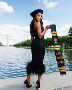 a woman in a black dress and hat is holding a scarf by the water's edge