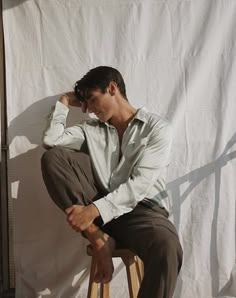 a man sitting on top of a wooden stool next to a white wall in front of a curtain