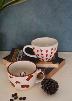 two coffee cups sitting on top of books next to a pine cone