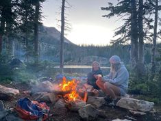 two people sitting around a campfire with backpacks on the ground next to them