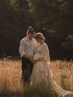 a pregnant woman and man standing in tall grass with trees in the background at sunset