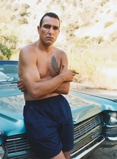 a shirtless man leaning on the hood of a car with his arms crossed in front of him