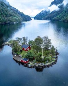 an island with two houses on it in the middle of a body of water surrounded by mountains