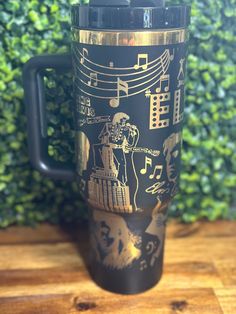 a black and gold travel mug sitting on top of a wooden table next to a green bush