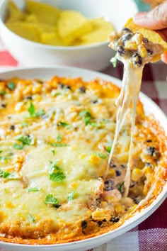 a hand holding a piece of pizza with cheese and toppings on it, over a casserole dish