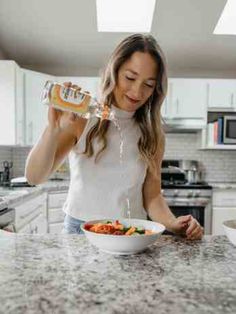 a woman standing at the kitchen counter drinking water from a bottle and eating food out of a bowl