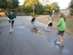 several children are playing in the street on scooters and skateboards with their parents