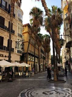 palm trees in the middle of a city street with people sitting at tables under umbrellas