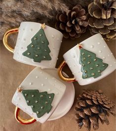 three coffee mugs with christmas trees on them and gold rings in front of pine cones