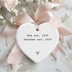 a white heart shaped ornament with pink ribbon and flowers in the background that reads mom ests 1970 grandma ests 2021