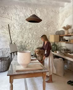 a woman standing in front of a wooden table holding a white vase and looking at it