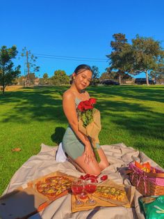 a woman sitting on a blanket in the grass with pizzas and flowers next to her