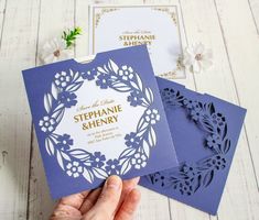 a person holding up two blue and white wedding cards with floral designs on the front