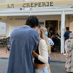 a man and woman standing in front of a building with people walking around it on the sidewalk