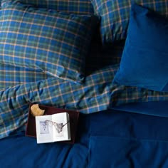 an open book on top of a bed with blue sheets and plaid pillowcases