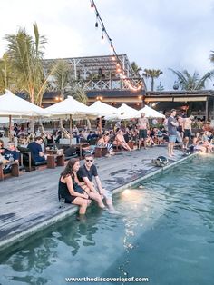 people sitting on the edge of a pool with umbrellas and tables in the background