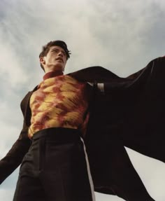 a man in a suit and tie with his arms spread out to the side while standing under a cloudy sky