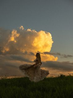 a woman in a long dress standing on top of a lush green field under a cloudy sky