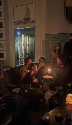 a group of people sitting around a cake with a candle on it in the middle of a living room