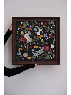 a person holding up a framed painting with flowers and leaves on it in front of a white wall